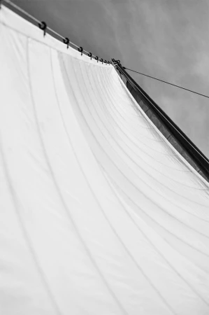 Monochrome photo of a sailboat sail with grommets along the mast, with a cloudy sky visible in the background on a sunny day.