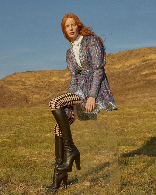 A red-haired woman in a patterned dress and boots sits on a clear box in a grassy field with a hill under a bright blue sky.
