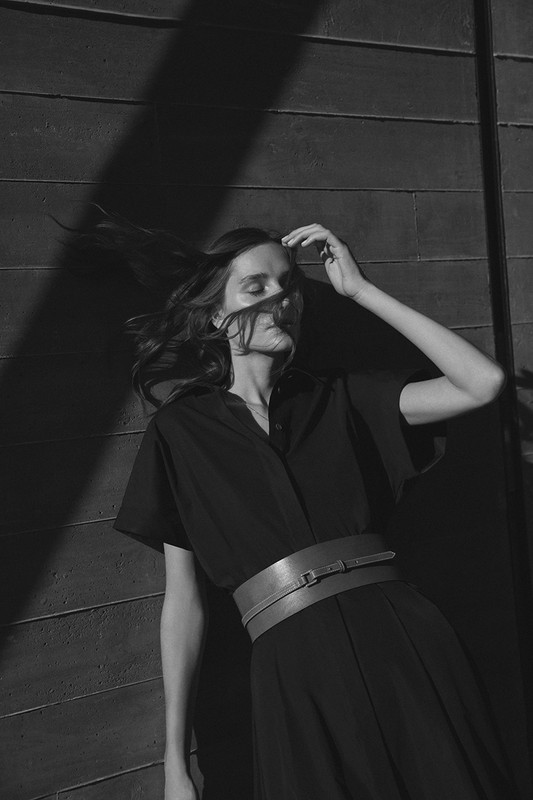 Monochrome portrait of a woman leaning against a dark paneled wall with hair blowing across her face and wearing a wide belt.