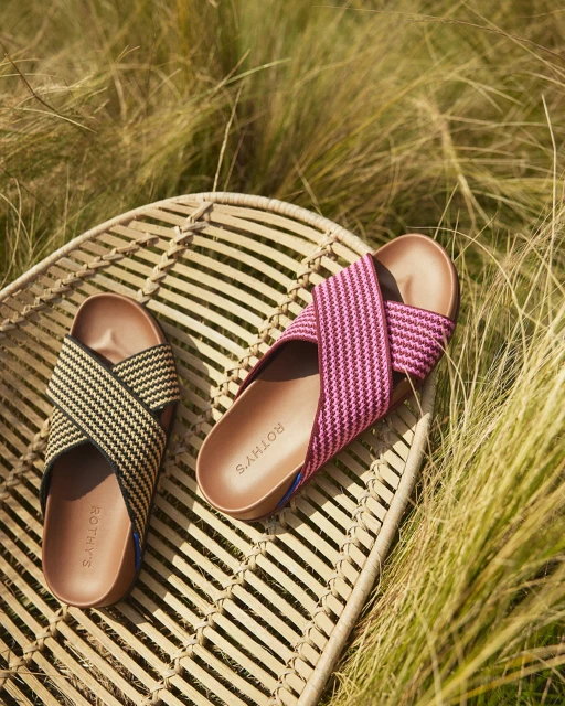 Close-up of Rothy's crisscross sandals in pink and black & beige on a woven table, outdoors in natural light with tall grass backdrop.