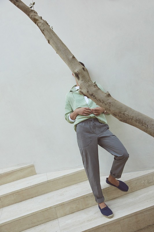 Man in green sweater, grey pants, and navy shoes leaning against a tree trunk on stairs. Minimalist fashion photography. 