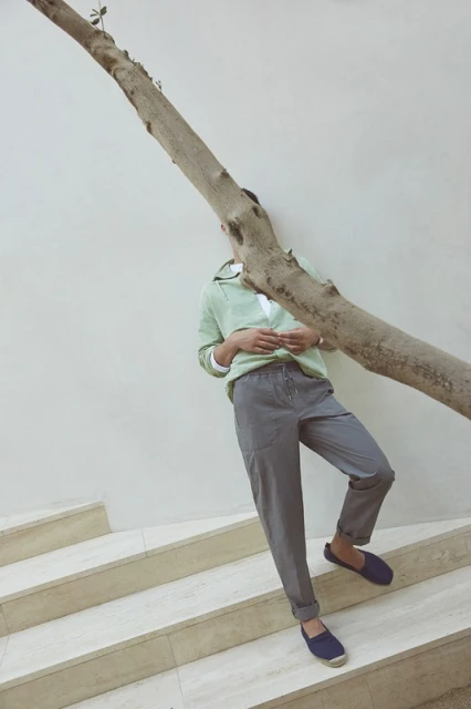 Man in green sweater, grey pants, and navy shoes leaning against a tree trunk on stairs. Minimalist fashion photography. 
