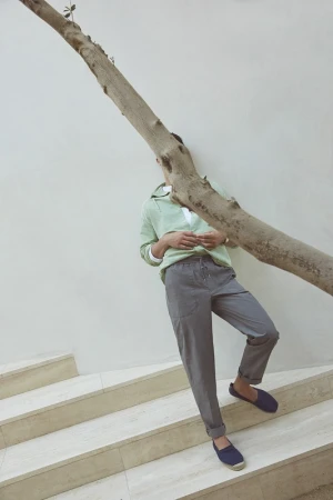 Man in green sweater, grey pants, and navy shoes leaning against a tree trunk on stairs. Minimalist fashion photography. 