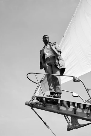Low angle shot of a man in a trench coat standing on the bow of a sailboat in black and white on a sunny day with a clear sky.