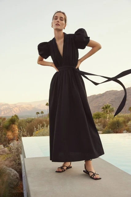 A woman models a black dress with a plunging neckline and puff sleeves, set against a backdrop of mountains and a swimming pool.