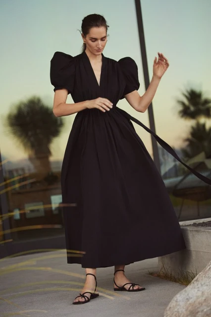 A woman wearing a black dress with puff sleeves and strappy sandals stands outdoors, reflecting light visible on a glass wall behind.