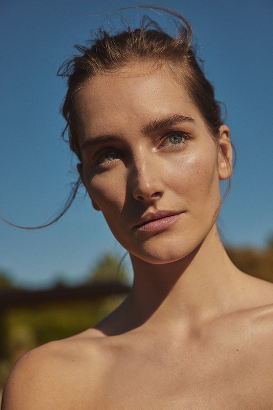 Close-up portrait of a woman with fair skin, fair hair pulled back, looking at the viewer against a blue sky and blurred greenery.
