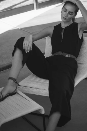 Monochrome portrait of a fair-skinned woman in a sleeveless black jumpsuit sitting on a white woven chair outdoors with neutral background.
