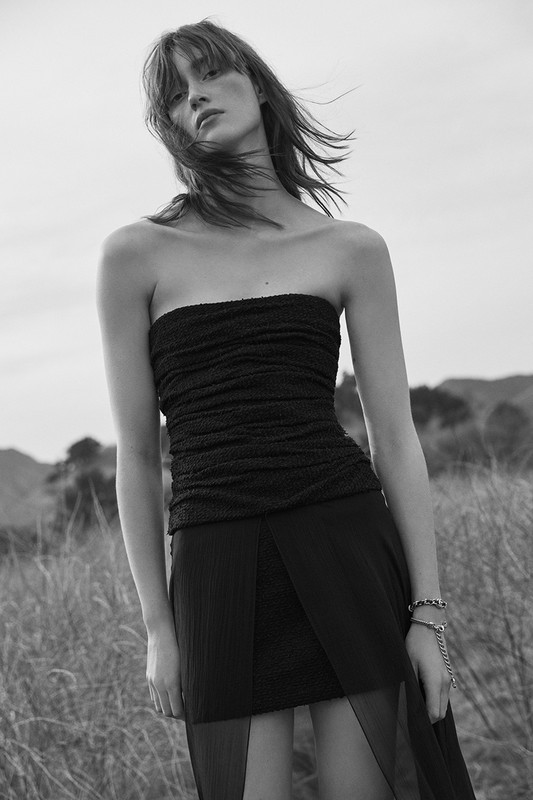 Monochrome portrait of a woman wearing a strapless dark top and skirt standing outdoors in dry tall grass.