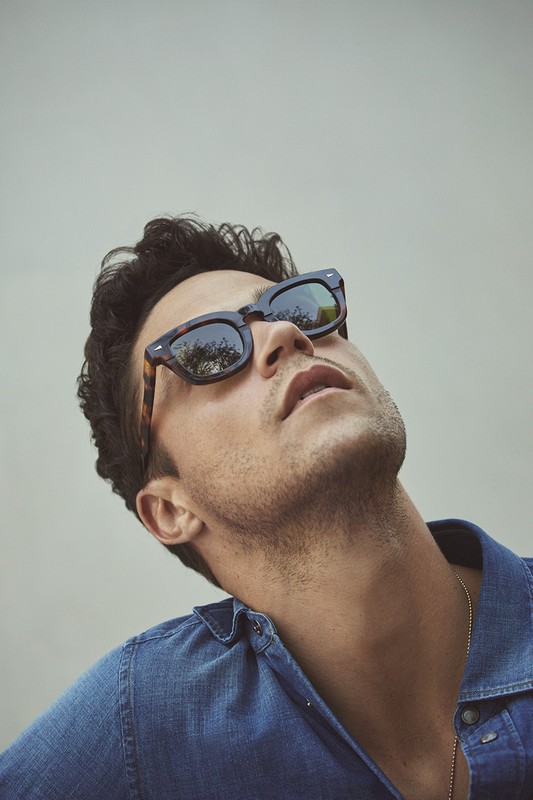 Adult male wearing sunglasses looks up in a low-angle shot. He is wearing a blue buttoned shirt and a gold chain against a grey backdrop.