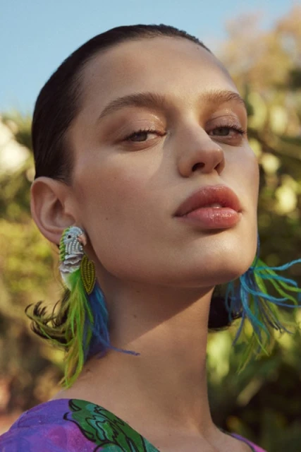 Portrait of a female model with light skin wearing earrings shaped like birds with blue and green feathers on an outdoor backdrop.