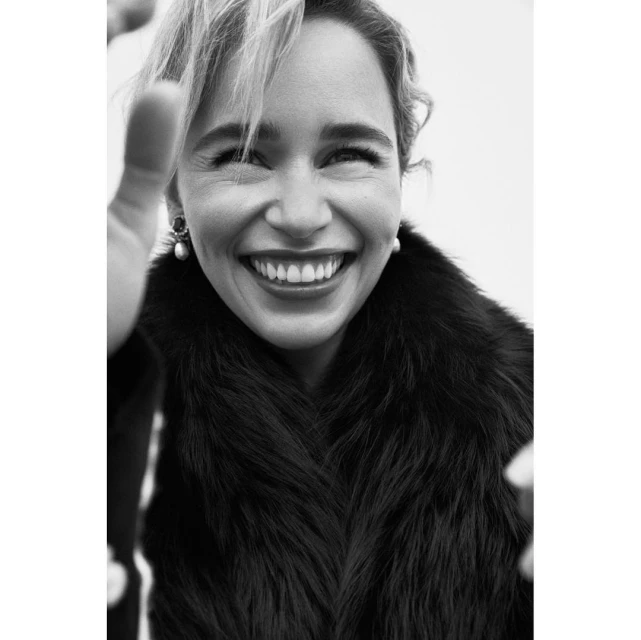 Black-and-white portrait of a smiling adult female with fair skin, wearing a fur coat and pearl earrings against a light background.