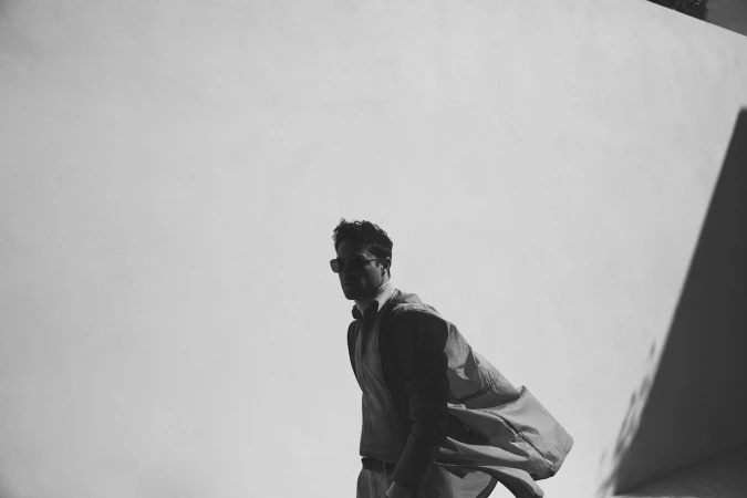 Man wearing sunglasses and a jacket standing against a bright wall in a black and white portrait.