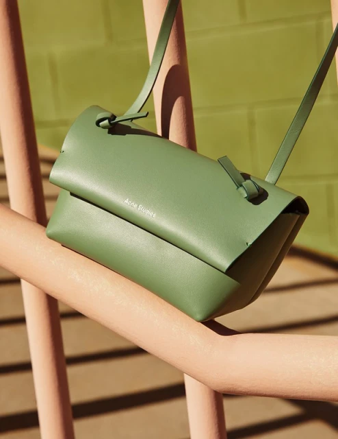 Close-up of a green Acne Studios leather handbag on a pink metal railing with an out-of-focus green wall background.