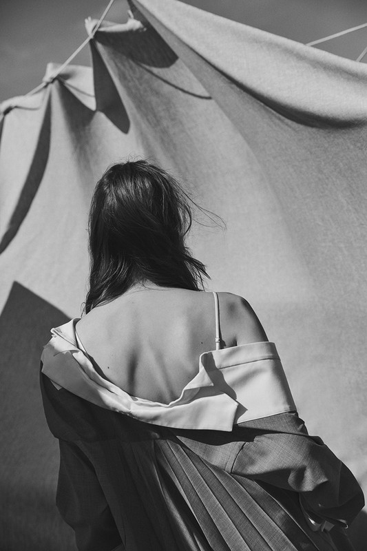 Back view of a woman in a coat with a draped cloth background, monochrome fashion, outdoor shot. Simple composition.