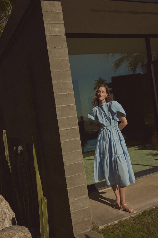 Model in a light blue tiered midi dress standing near a modern concrete building with large glass windows and palm trees reflected.
