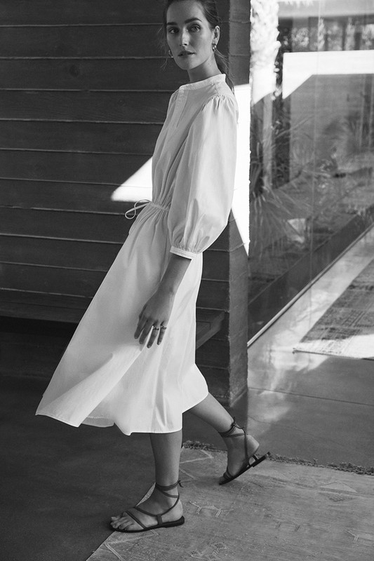 Monochrome shot of a woman in a white dress and strappy sandals walking on a concrete floor, with a wooden wall in the background.