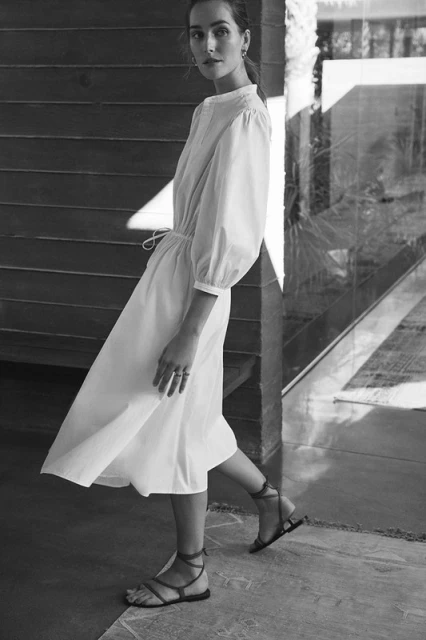 Monochrome shot of a woman in a white dress and strappy sandals walking on a concrete floor, with a wooden wall in the background.