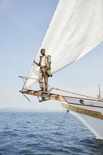 Man on a sailboat bow in beige jacket, brown pants, and white shirt against blue sky and ocean water, sailing on a sunny day.