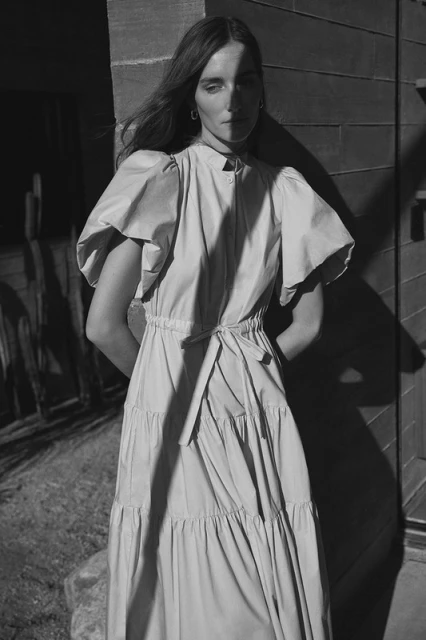 Monochrome outdoor portrait of a stylish woman in a tiered maxi dress with puff sleeves, standing in front of a dark wood-paneled building.