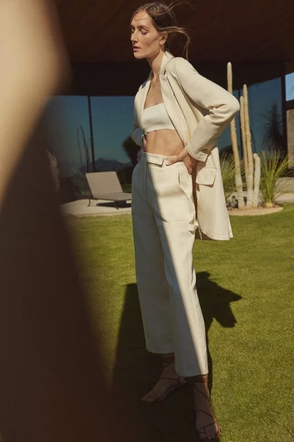 A stylish adult female in a light-colored two-piece suit poses on a sunny lawn with modern architecture and cacti in the background.