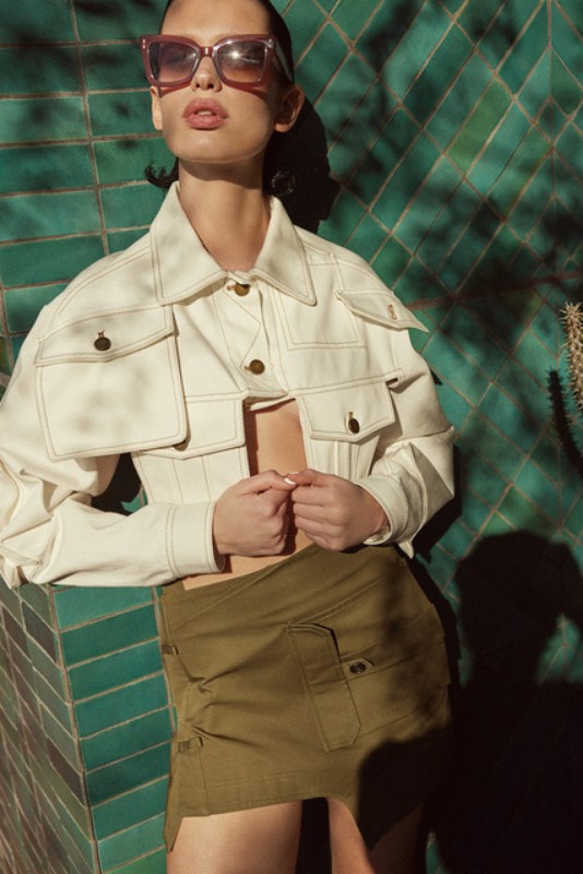 A woman poses in a white jacket, olive skirt, and pink sunglasses against a green tiled wall with dappled shadows creating a fashion portrait.