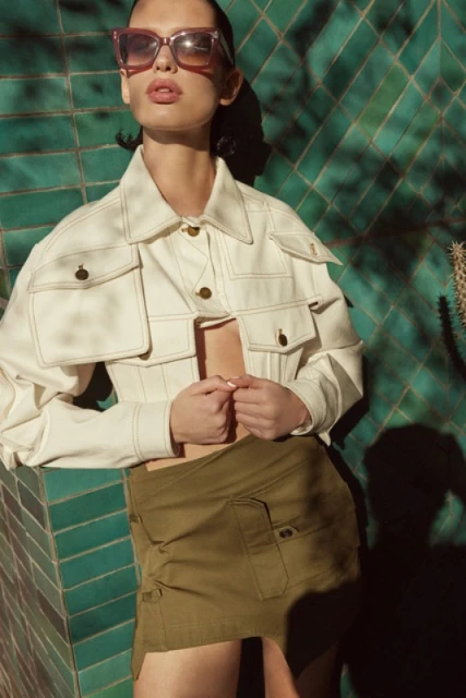 A woman poses in a white jacket, olive skirt, and pink sunglasses against a green tiled wall with dappled shadows creating a fashion portrait.