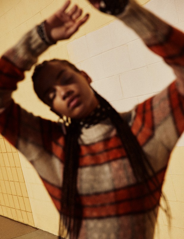 Fashion portrait of a woman with her arms raised, wearing a striped sweater with an orange pattern against a neutral background.