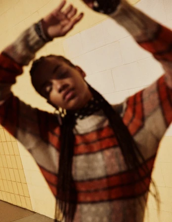 Fashion portrait of a woman with her arms raised, wearing a striped sweater with an orange pattern against a neutral background.