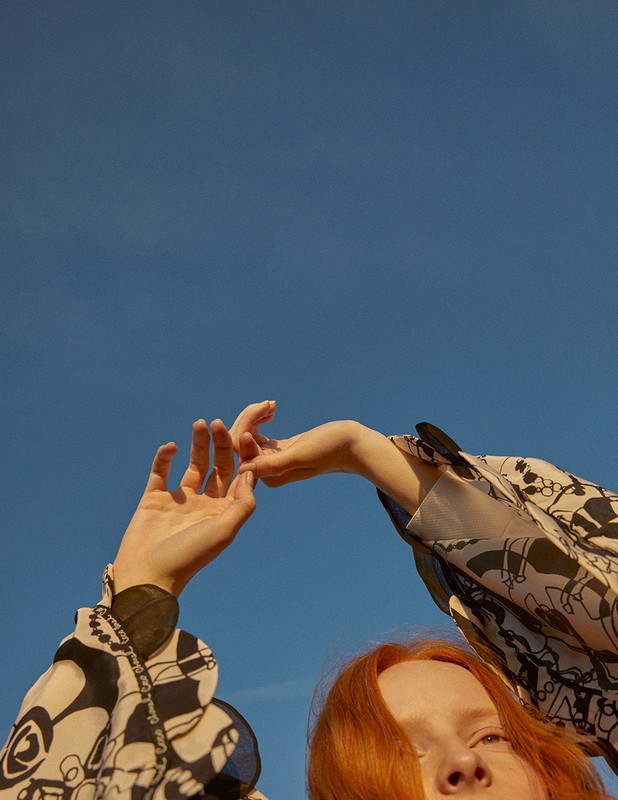Woman with red hair poses with arms raised against a blue sky wearing a patterned long sleeve shirt in an outdoor setting.