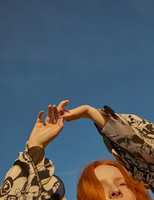 Woman with red hair poses with arms raised against a blue sky wearing a patterned long sleeve shirt in an outdoor setting.