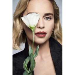 Close-up portrait of Emilia Clarke holding a white flower covering one eye against a neutral background.