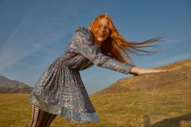 Red-haired woman in a patterned metallic dress leaning on a clear glass block in an arid mountainous landscape under a blue sky.