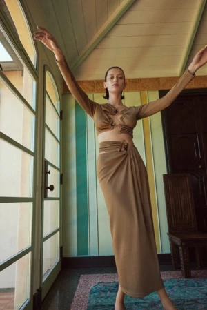 Model in brown two-piece outfit with arms raised inside a room with arched glass doors and green striped walls