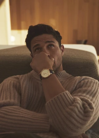 A man in a ribbed knit sweater wearing a wristwatch, posing indoors with a warm, soft-lit background.