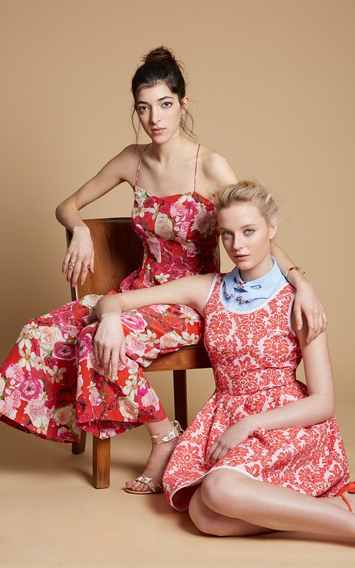 Two female models in red patterned dresses posing together with a wooden chair in a studio setting