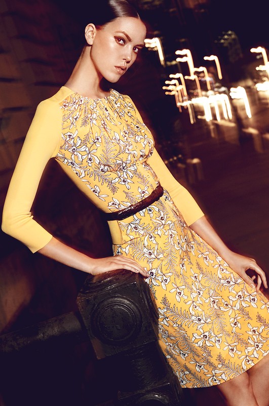 A fashionable woman in a yellow floral dress poses against an urban backdrop, showcasing her attire and sophisticated style.