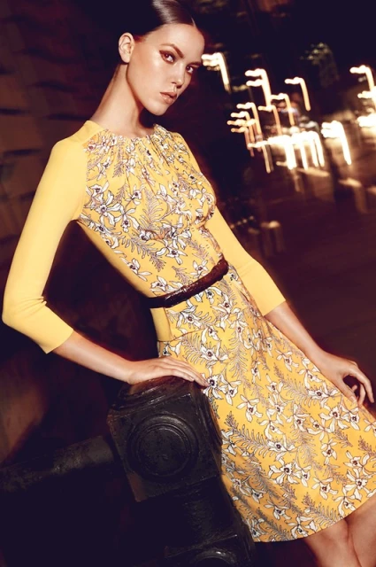 A fashionable woman in a yellow floral dress poses against an urban backdrop, showcasing her attire and sophisticated style.