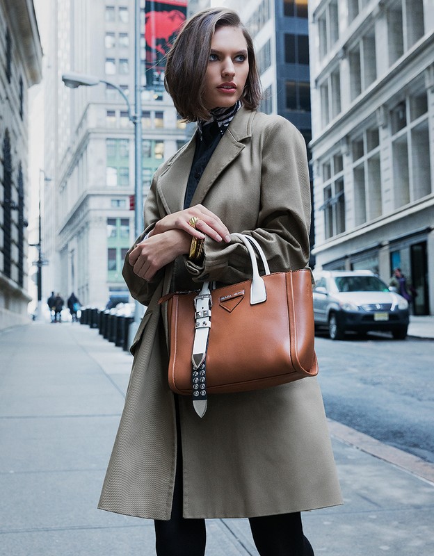 Woman with short dark hair wearing a tan coat carrying a brown leather satchel in a city street with buildings and cars.