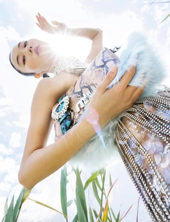Eye-level shot of an adult woman in a snakeskin-patterned dress, holding a matching purse against a bright sky and green plants.