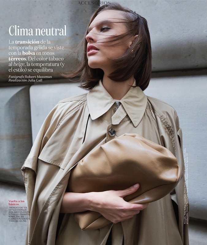 Fashion editorial shot of a woman holding a tan purse while wearing a beige trench coat, posing against a concrete wall.