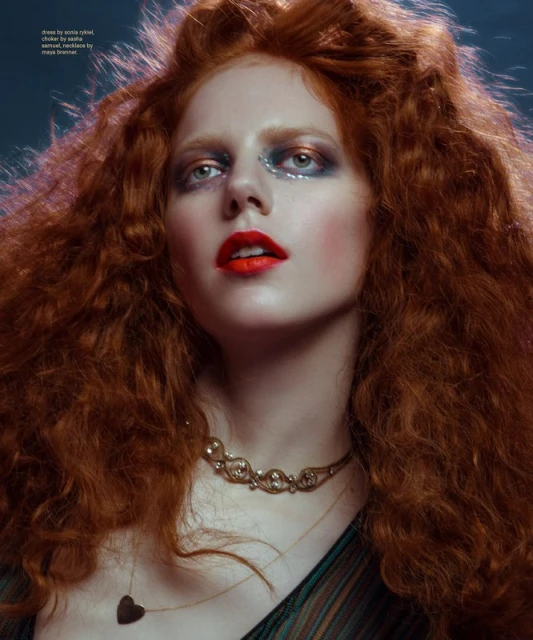 Close-up portrait of a red-haired woman with elaborate makeup and jewelry, facing upwards in a dimly lit, hazy environment.