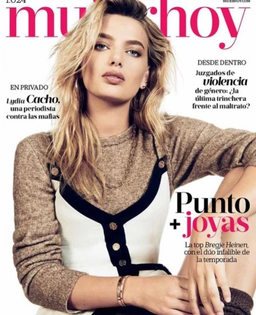 Model Bregje Heinen posing for a magazine cover wearing a brown sweater and a white dress with black trim against a white background.