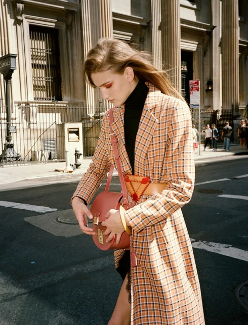 A woman wearing a plaid coat and holding a purse walks across the street in a city with a building in the background.