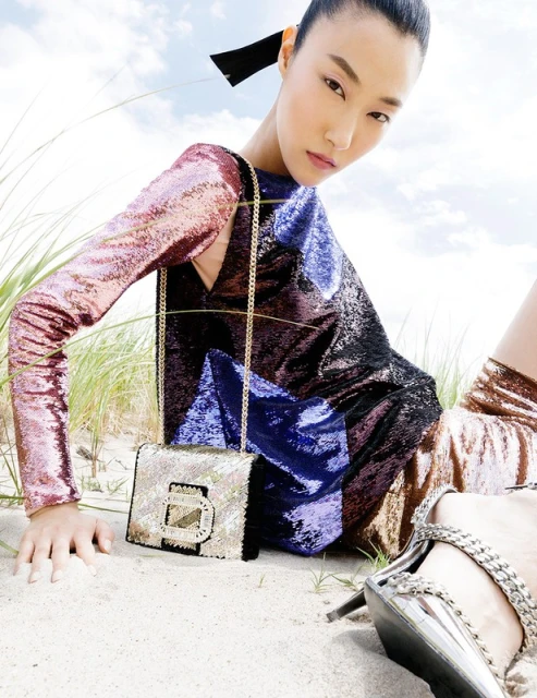 Asian woman poses in a multicolored sequin dress with a handbag on a sandy beach with dune grass and a bright sky backdrop.