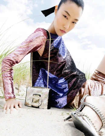 Asian woman poses in a multicolored sequin dress with a handbag on a sandy beach with dune grass and a bright sky backdrop.