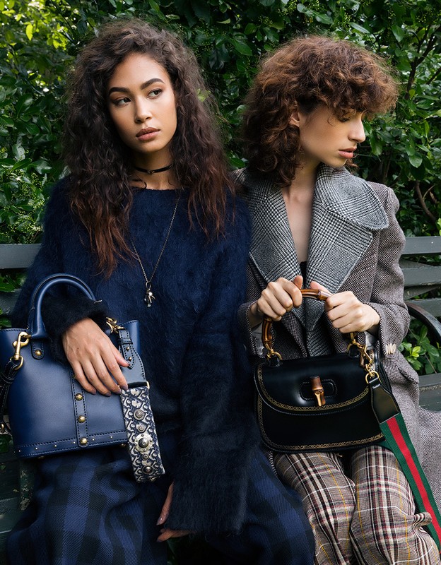 Two women pose with purses in a park. The woman on the left wears a blue sweater and skirt, while the woman on the right wears a grey coat.