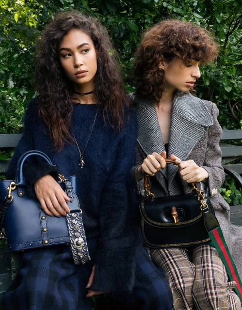 Two women pose with purses in a park. The woman on the left wears a blue sweater and skirt, while the woman on the right wears a grey coat.