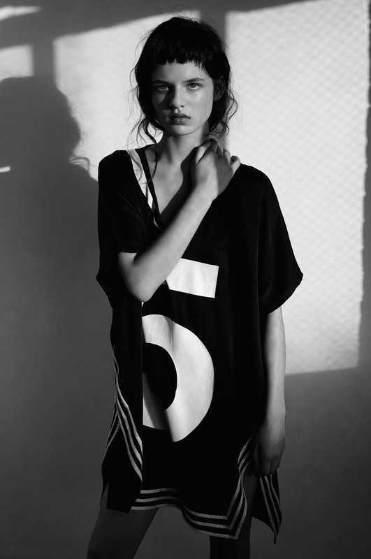 Monochrome portrait of a young woman with bangs in a black oversized shirt with white stripes and the number 5, against a light wall.