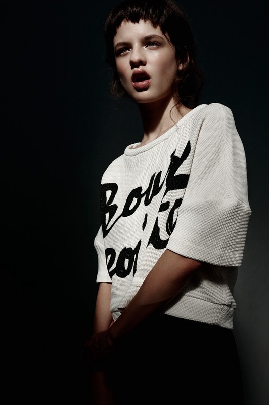 A young adult female model with dark hair and bangs wears a white printed sweater in a fashion shot against a dark background.